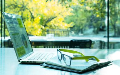 A laptop, notebook, and glasses resting on a tidy desk with a window view of healthy trees