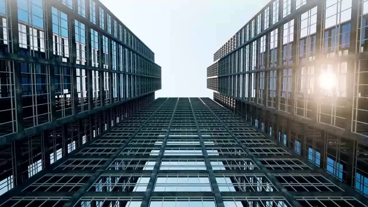 Upward view of two modern glass office towers with sunlight reflecting across the façades, symbolizing growth, leadership, and advancement; banner for Hogan Lovells’ 2026 partner and counsel promotions.