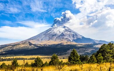 Mexico's Popocatepetl volcano surrounded by mountains, clouds against a bright blue sky, and trees in a yellow field